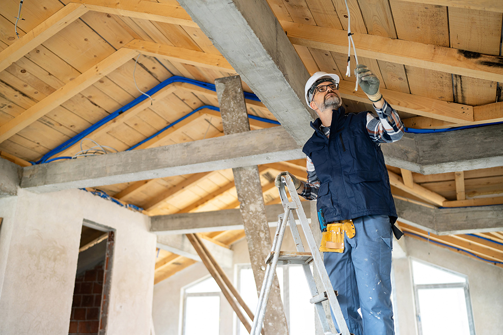 An electrician examines electrical wiring in a roof structure with wooden beams and concrete supports. He stands on a ladder, wearing protective gear, ensuring safety and compliance with building standards.